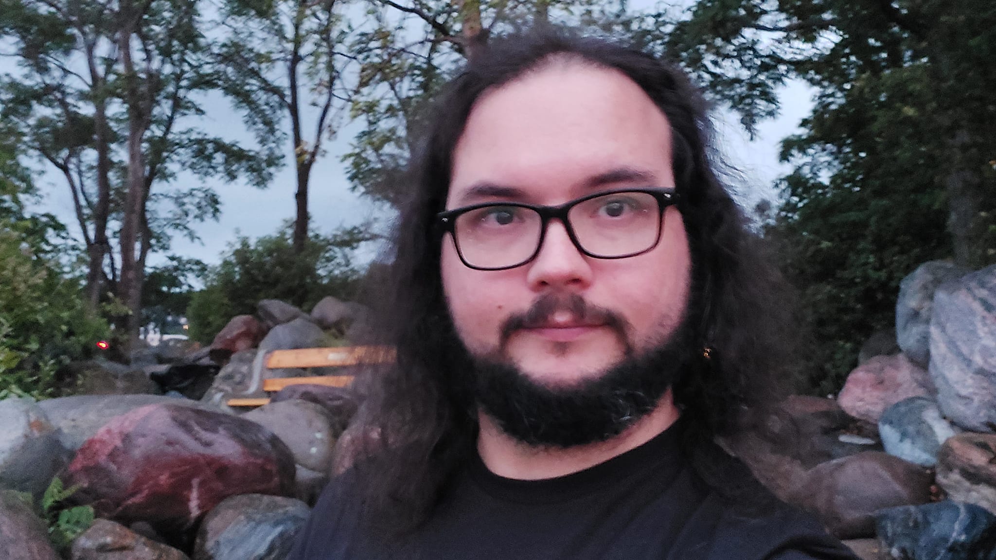 A man with long dark hair and a beard is at a sandy beach with a granite boulder breakwater, photo 1